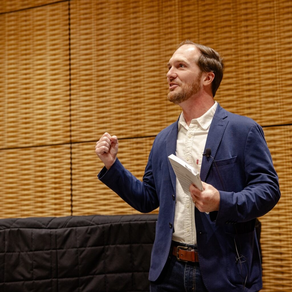 Photo of Dustin M. Hoffman holding a book and gesturing while speaking